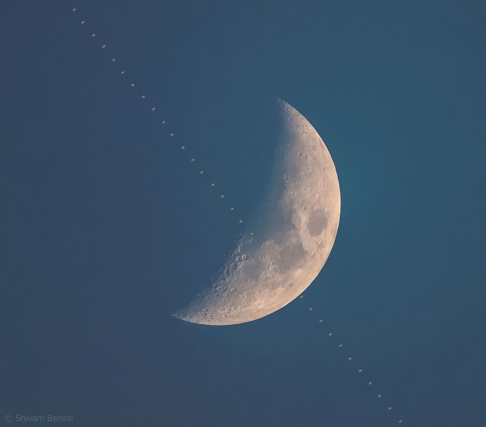 Crescent Moon in a blue evening sky with a diagonal line of small dots, a stacked sequence showing the ISS transiting past it.