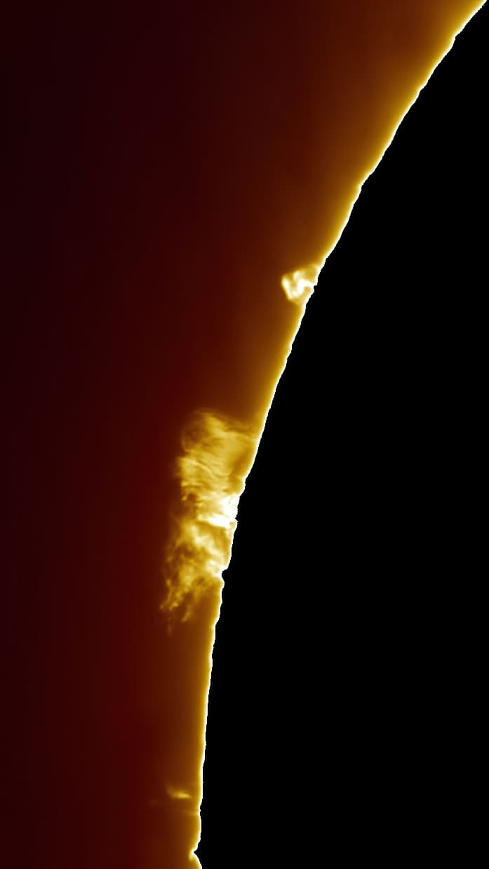 Close-up H-alpha image of a tall, bright solar prominence jetting upward from the sun's limb against a dark red chromosphere background.